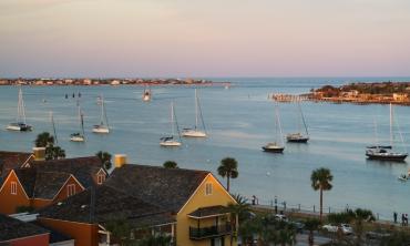 A rooftop view of the St. Augustine Harbor, showing brightly painted buildings with the bay with many sailboats on moorings beyond, a shrimp boat returing to harbor, and the St. Augustine inlet to the Atlantic