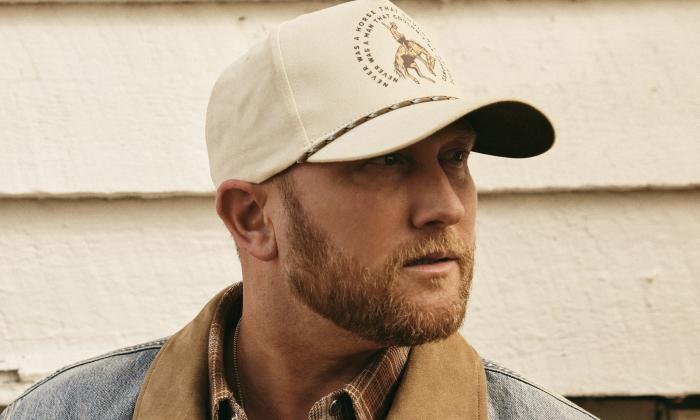 Cole Swindell, sporting a cap, poses against an off-white backdrop.