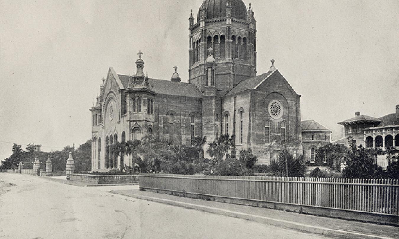 A black and white photograph of a grand church in St. Augustine, Florida. In the foreground is a sandy street and a small pile of horse droppings. A fence extends from the right hand side of the image towards the focus of the photo, which is a massive church. The church is made of stone and stands at well over three stories tall, the tallest structure of the church being a domed tower that has a cross on top. Palm trees being grown on the ground are dwarfed by the beautiful building's spires and windows.