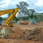 A full color image of a yellow bulldozer in a cleared landscape surrounded in piles of dirt. There are trees and a few other colorful machines in the background.