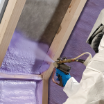 A full color image of a contractor in full protectiv gear spraying purple foam insulation between two beams.